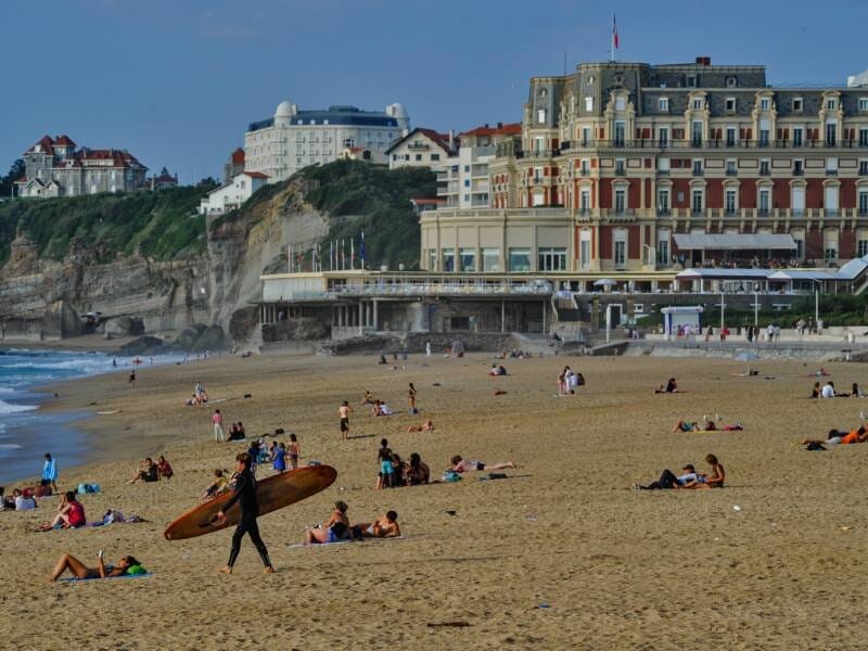 Plage animée avec des baigneurs, un surfeur portant sa planche et des bâtiments anciens en arrière-plan.