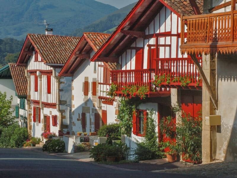 Maisons traditionnelles blanches avec volets rouges et balcons fleuris dans un village au pied des montagnes.