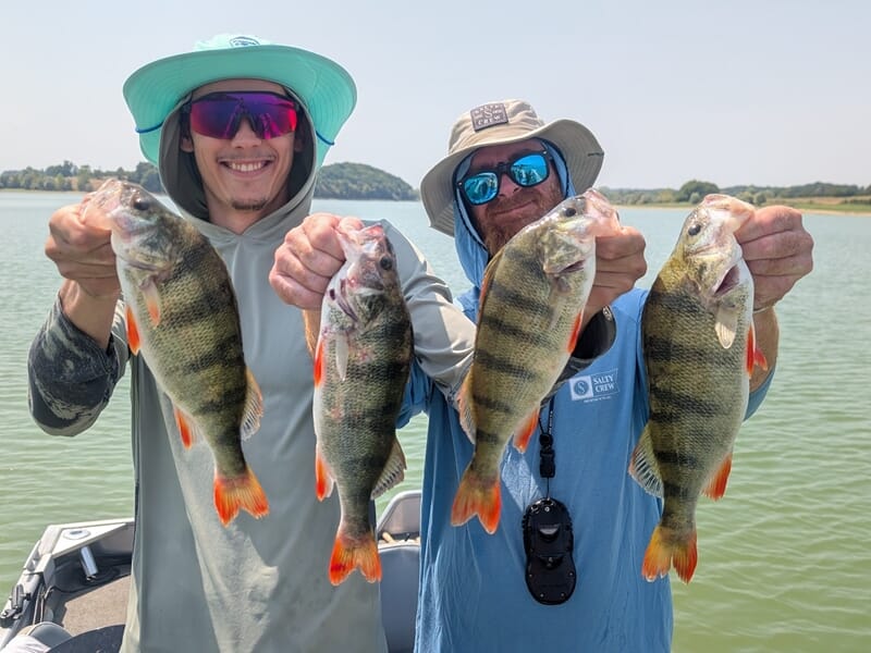 Deux pêcheurs souriants tenant chacun deux perches colorées au bord d'un lac sous un ciel clair.