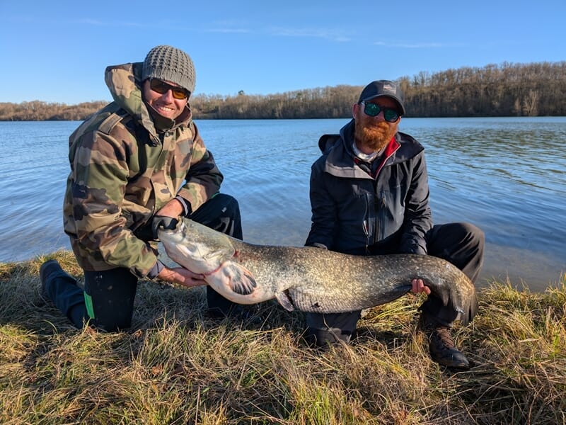 Deux hommes assis sur l'herbe près d'un lac, tenant un très gros poisson lors d'un stage de pêche à Toulouse.
