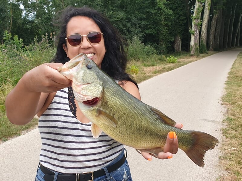 Femme souriante en débardeur rayé tenant un gros poisson noir et vert lors d'un stage de pêche à Toulouse.