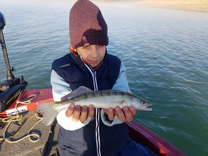 Un garçon en bonnet et gilet bleu tient un poisson fraîchement pêché sur un bateau au bord de l'eau calme.