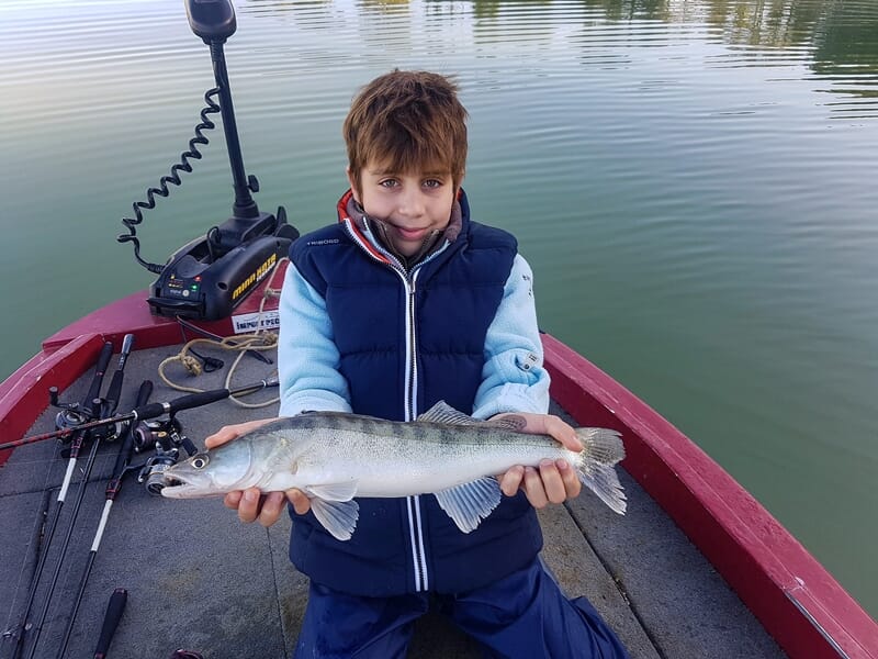 Un enfant souriant en gilet bleu présente un poisson long et argenté sur un bateau rouge au milieu d'un lac.