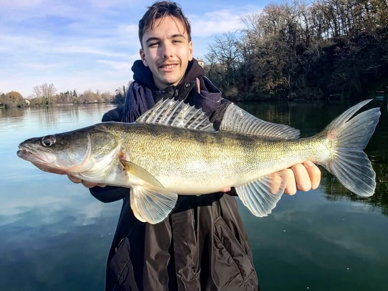 Un jeune homme en manteau noir montre fièrement un gros poisson doré devant un paysage d'eau et d'arbres.