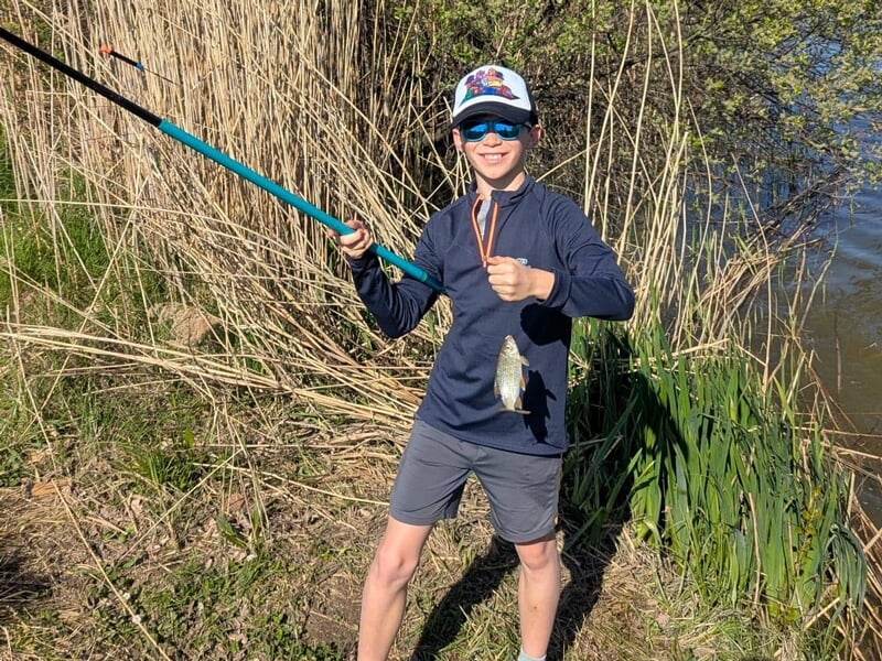 Un garçon en tenue sportive et casquette tient une canne à pêche avec un petit poisson accroché au bout.