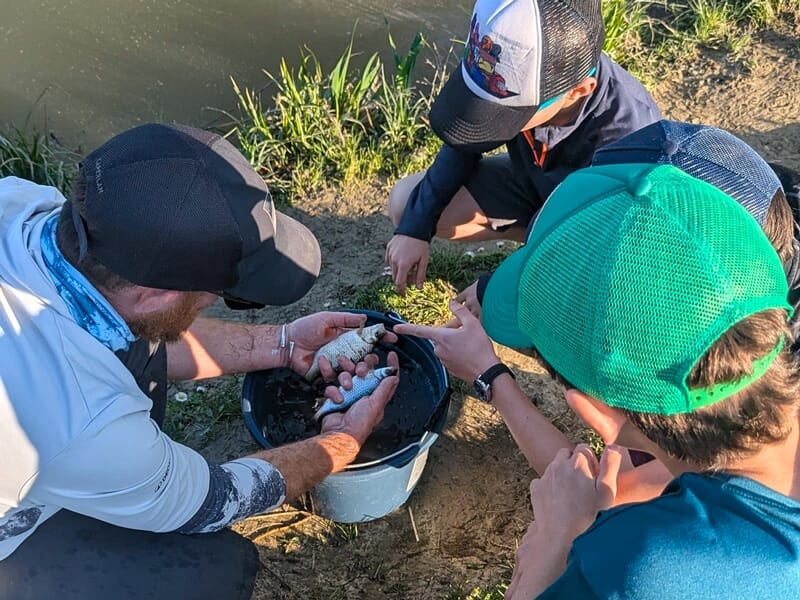 Un groupe de jeunes observe un moniteur qui montre un poisson dans un seau lors d'une activité de pêche.