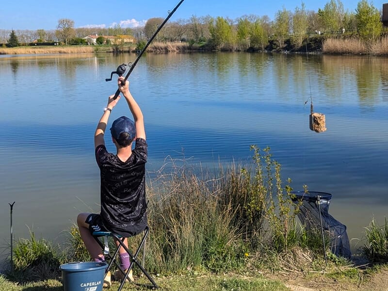 Un pêcheur assis au bord d'un étang lance sa ligne dans l'eau entourée de végétation et d'herbes hautes.