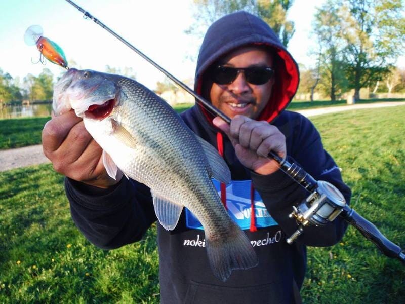 Homme souriant en sweat à capuche tenant une canne à pêche et un poisson fraîchement pêché lors d'un stage de pêche dans l'Ain.