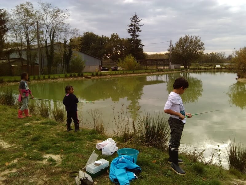 Trois enfants pêchant au bord d'un étang calme entouré d'herbe et d'arbres lors d'un stage de pêche dans l'Ain.