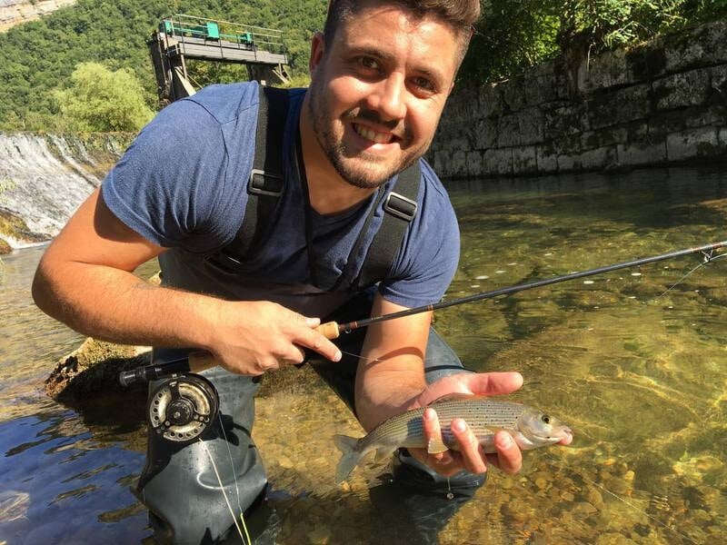 Jeune homme accroupi dans une rivière claire, tenant une truite et une canne à pêche lors d'un stage de pêche dans l'Ain.