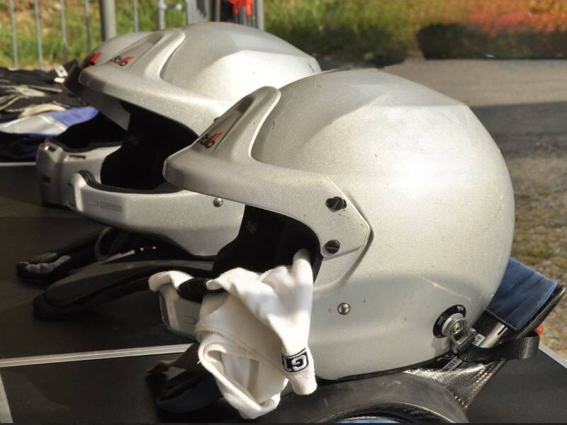 Deux casques de pilote argentés posés sur une table avec une paire de gants blancs, ambiance circuit automobile.