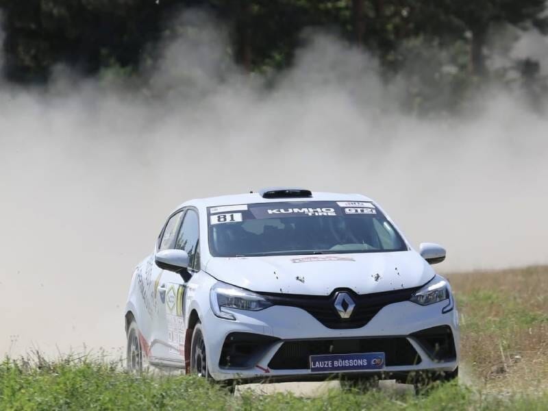 Voiture de course blanche avec logos, roulant sur une piste poussiéreuse entourée d'herbe et d'arbres.