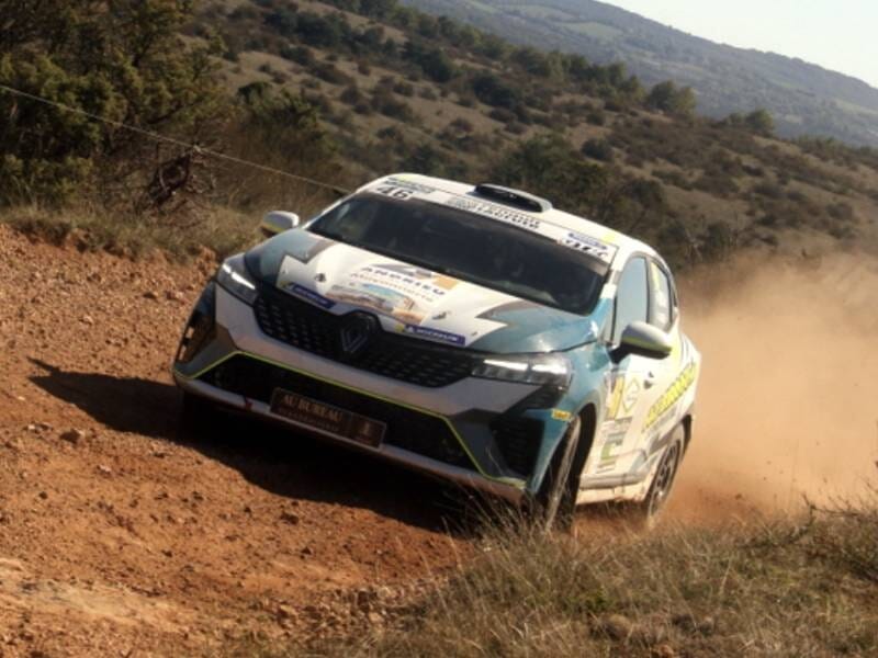 Voiture de rallye Renault blanche et bleue dérapant sur piste terreuse avec poussière, paysage vallonné en arrière-plan.