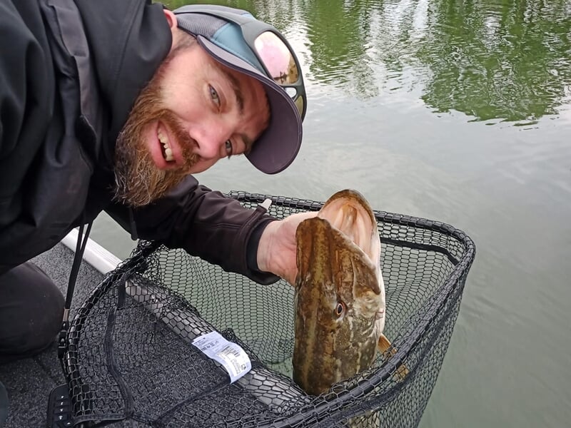 Homme souriant tenant un poisson dans un filet au bord d'un plan d'eau calme lors d'un stage de pêche près de Dijon.