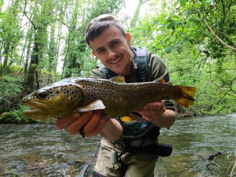 Un homme souriant tient une grosse truite fario au bord d'une rivière entourée d'arbres verts en arrière-plan.