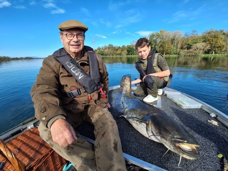 Lors d'un stage de pêche près de Colmar, un homme âgé et un garçon, posent sur un bateau avec un grand poisson plat sur un lac calme sous un ciel bleu.