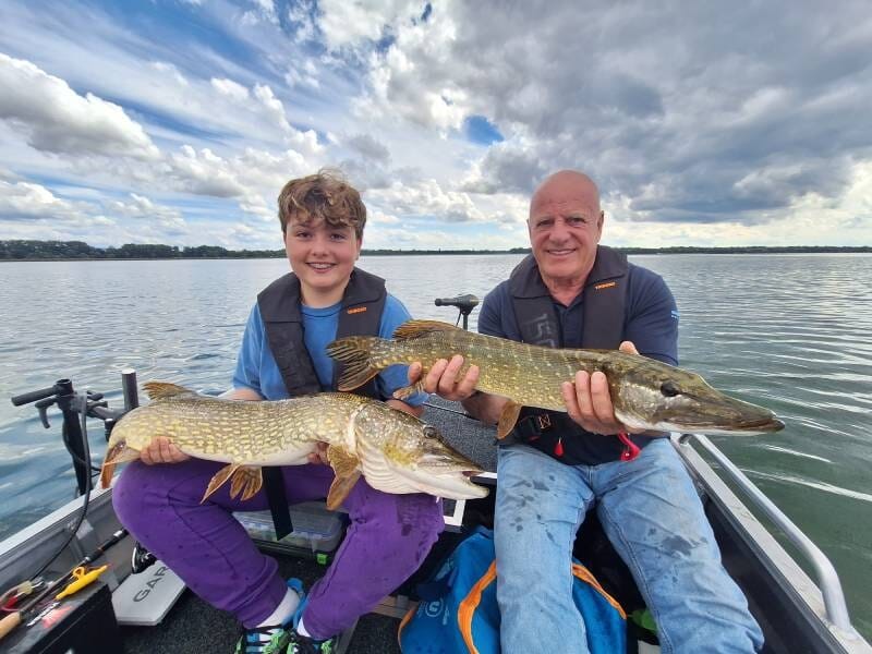 Un homme et un garçon assis sur un bateau montrent fièrement deux grands brochets capturés sur un lac sous un ciel nuageux.