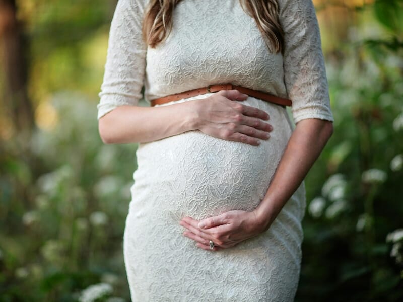 Femme enceinte en robe blanche dentelle, posant dans un jardin verdoyant lors d'un shooting photo grossesse à Dunkerque.