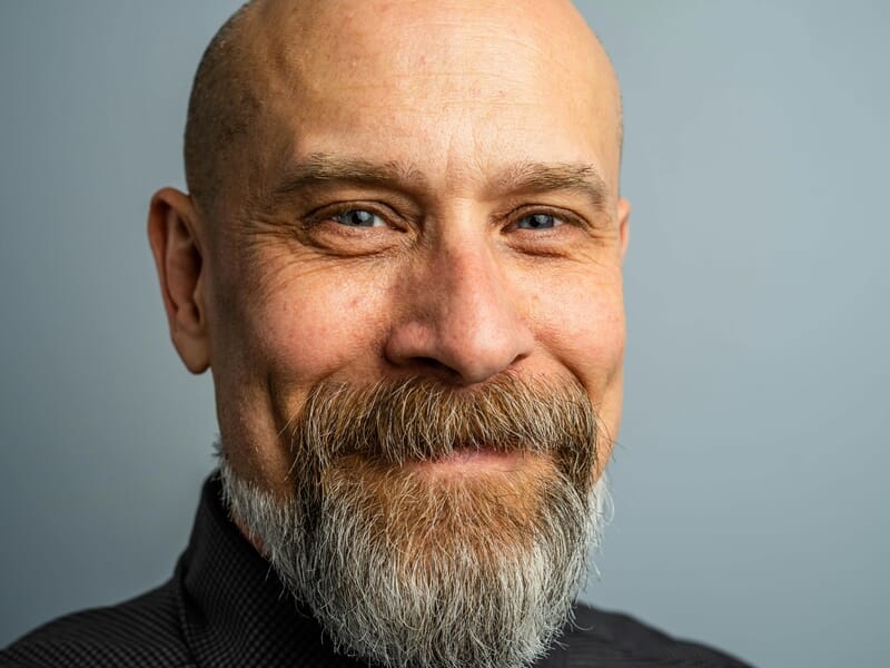 Gros plan d'un homme chauve avec barbe grise souriant, fond bleu clair lors d'un shooting photo portrait à Dunkerque.