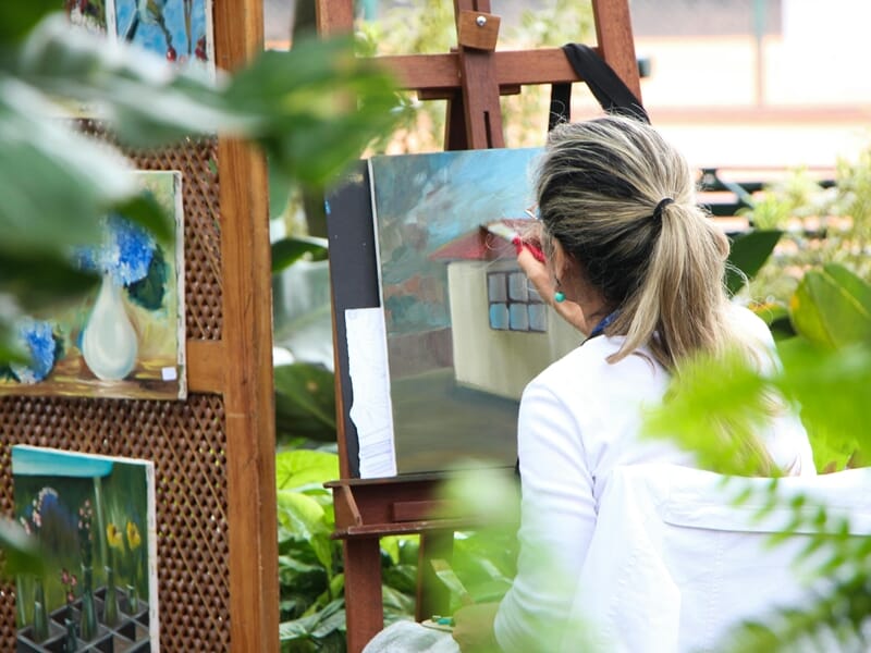 Une femme peint sur une toile dans un atelier lumineux entouré de plantes lors d'un shooting photo lifestyle à Dunkerque.