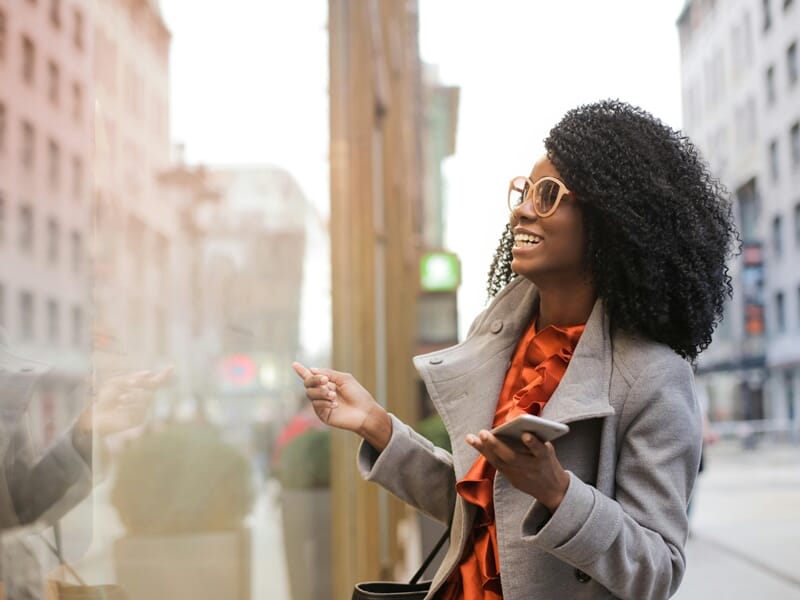 Une femme souriante regarde une vitrine en ville, tenant un smartphone, lors d'un shooting photo lifestyle à Dunkerque.