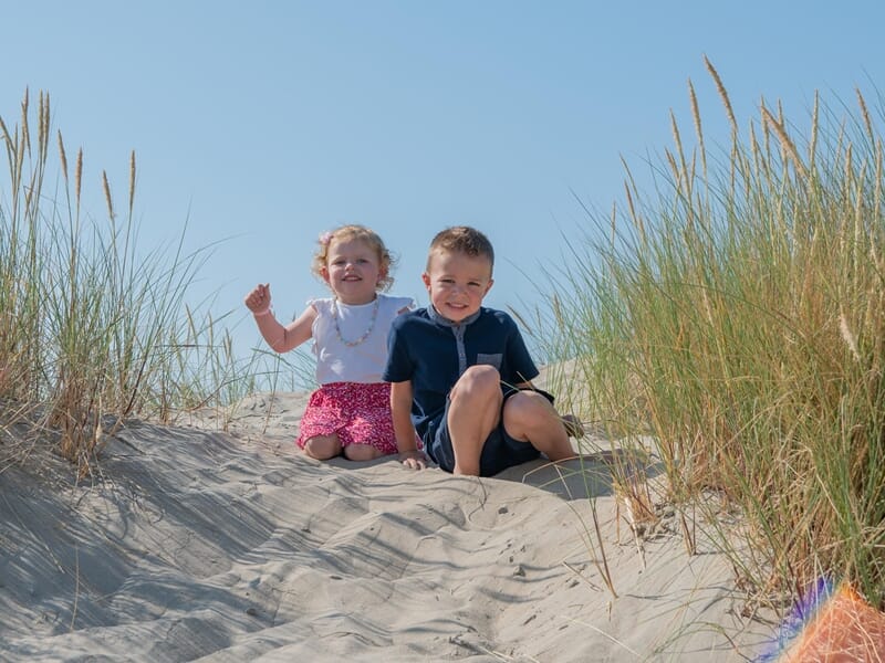 Deux enfants assis sur une dune de sable avec des herbes, sous un ciel bleu clair ensoleillé lors d'un shooting photo famille à Dunkerque.