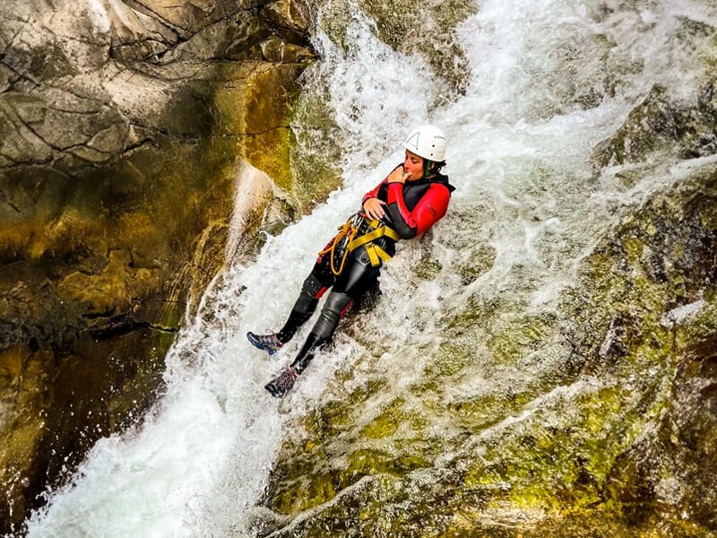 Personne en tenue de canyoning glissant sur une cascade rocheuse avec casque et combinaison rouge et noire.