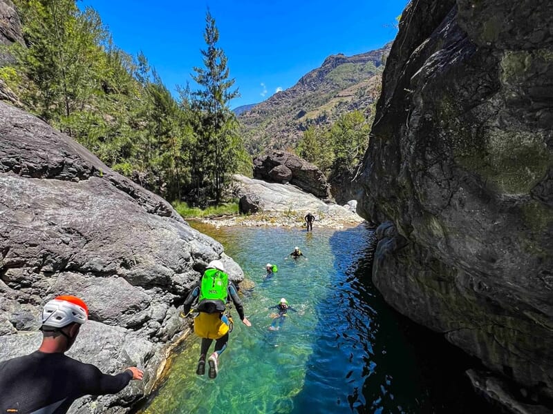 Groupe de personnes en combinaison et casque sautant dans une eau claire entourée de rochers et montagnes.