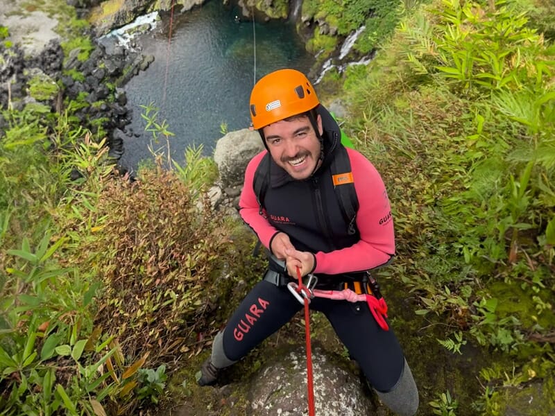 Homme équipé d'un casque orange et d'une combinaison de canyoning souriant en haut d'une falaise verdoyante.