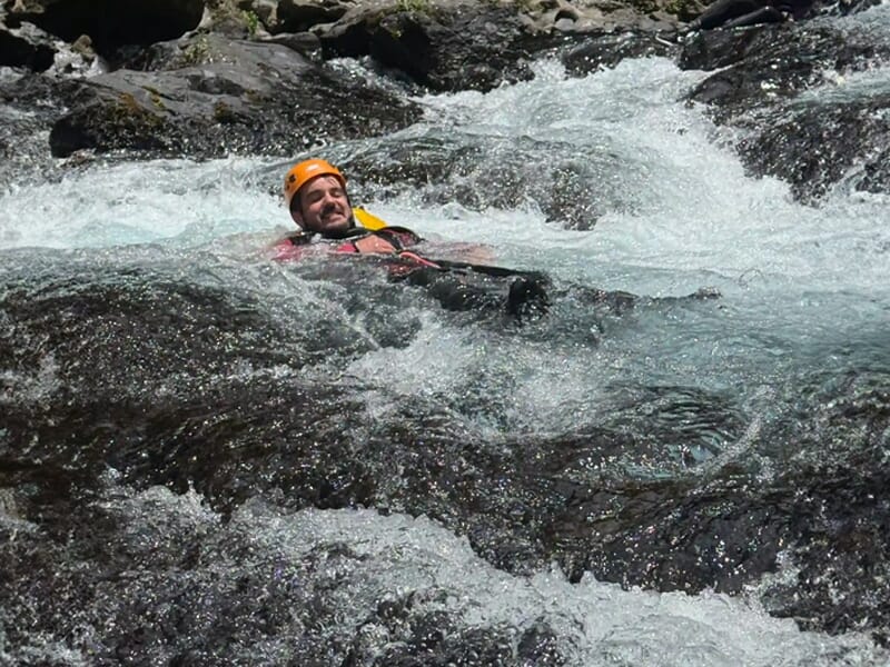 Homme en casque orange glissant dans un courant d'eau vive avec des rochers autour, souriant.
