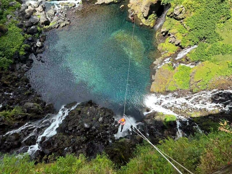 Vue aérienne d'une personne en canyoning descendant une cascade avec un casque orange et une corde.