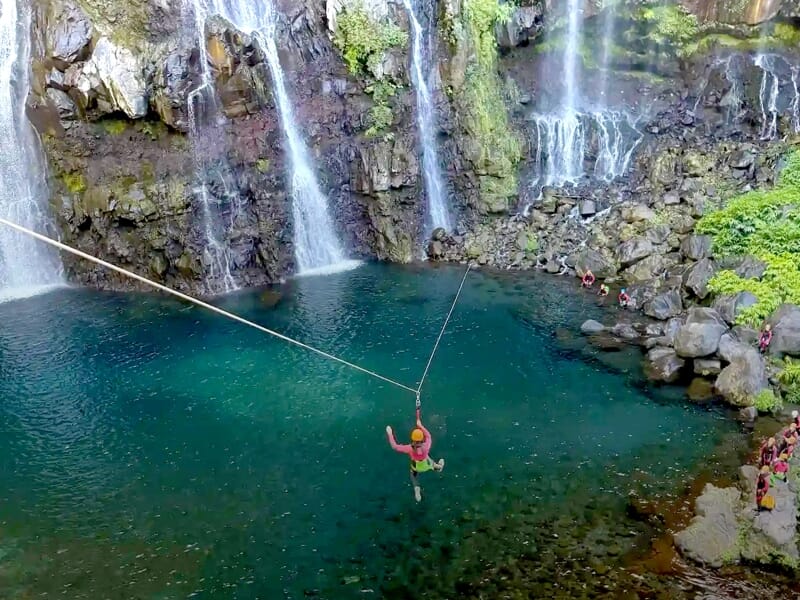 Personne suspendue à une tyrolienne au-dessus d'un bassin naturel entouré de cascades et de rochers à La Plaine des Palmistes.