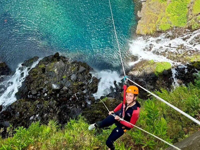 Femme souriante en équipement de canyoning sur une tyrolienne au-dessus d'une cascade et d'une végétation luxuriante.
