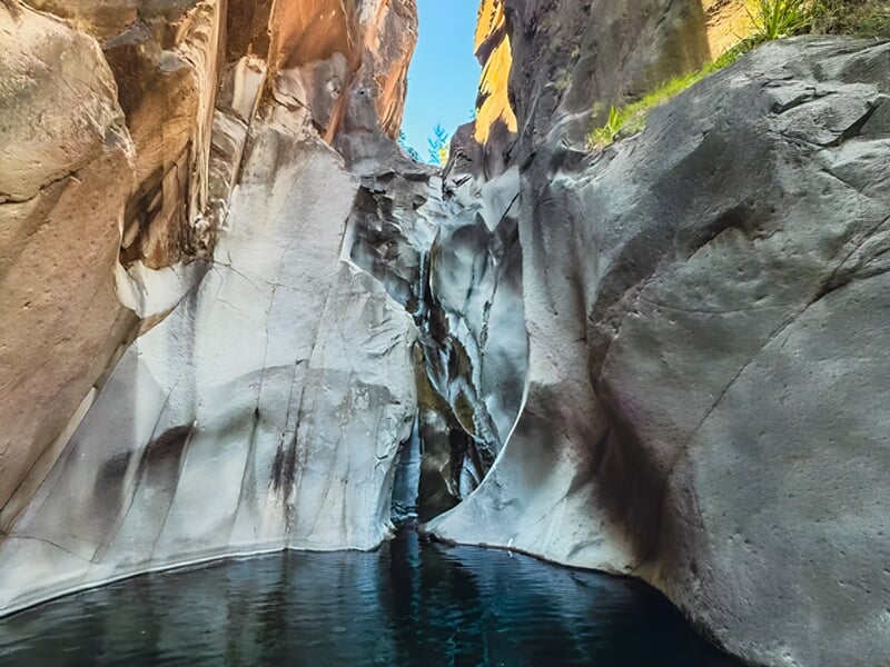 Canyon étroit avec eau calme et parois rocheuses lisses sous un ciel bleu clair ensoleillé.