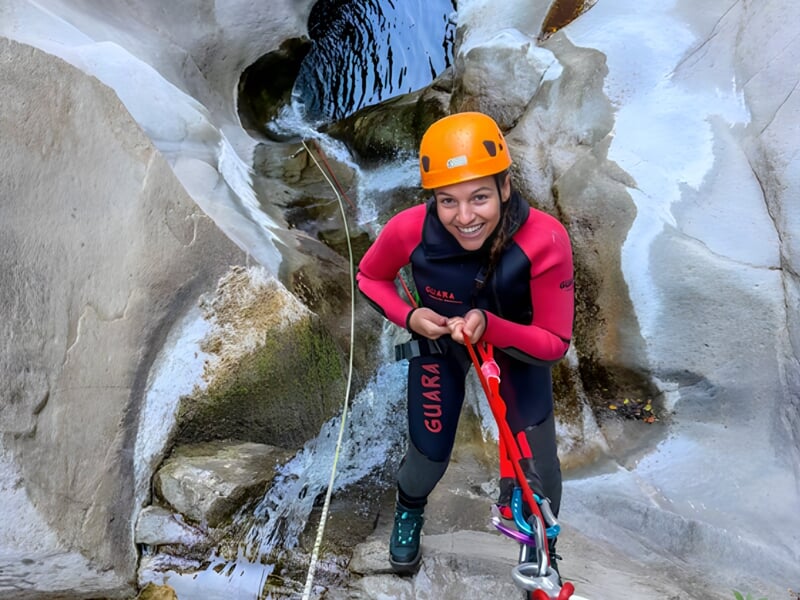 Femme souriante en tenue de canyoning avec casque orange et harnais, prête à descendre une cascade.