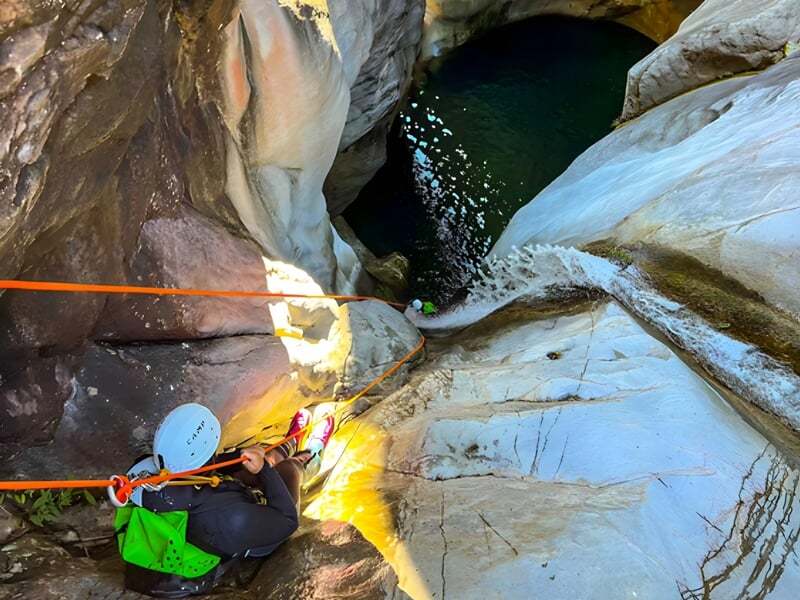 Personne en équipement de canyoning descend en rappel le long d'une paroi rocheuse blanche au-dessus d'un bassin d'eau.