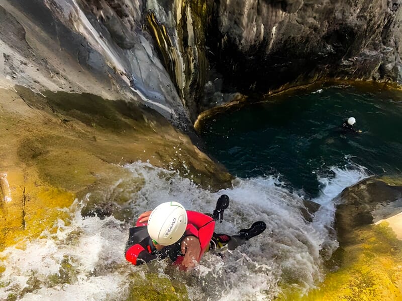 Canyoneur glissant sur une cascade rocheuse mouillée avec casque blanc et combinaison noire et rouge.