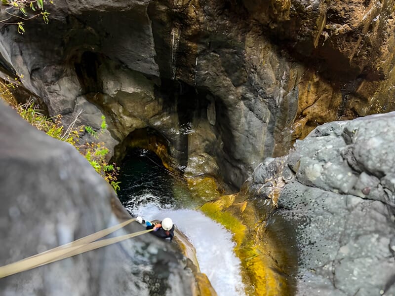 Vue plongeante sur un canyon avec une personne en rappel au-dessus d'une cascade et d'un bassin d'eau.