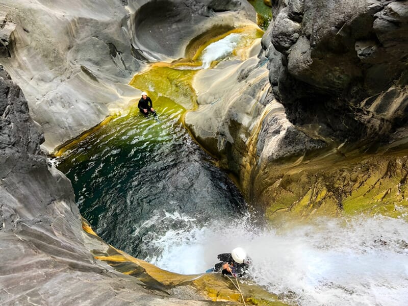 Deux personnes en combinaison de canyoning progressent dans un canyon rocheux avec eau et cascades.