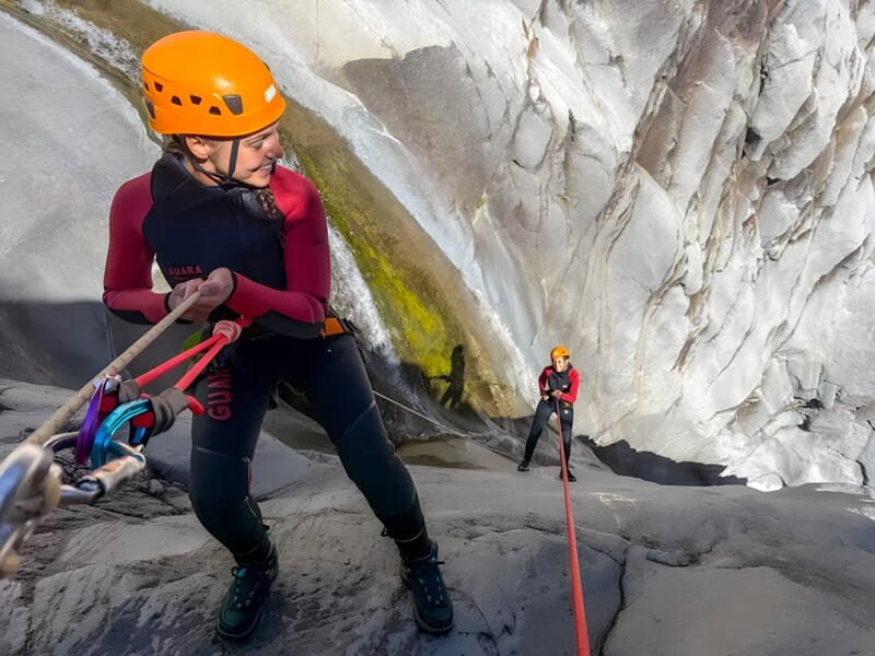 Femme en tenue de canyoning descend en rappel sur une paroi rocheuse blanche avec une autre personne en arrière-plan.