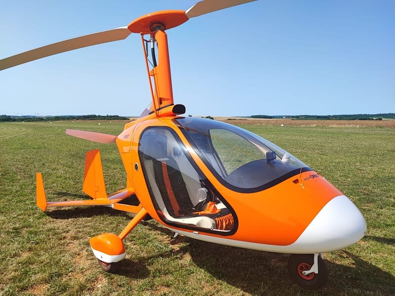 Un ULM autogire orange et blanc stationné sur un terrain d'herbe verte lors d'un baptême en ulm autogire dans le Cotentin.