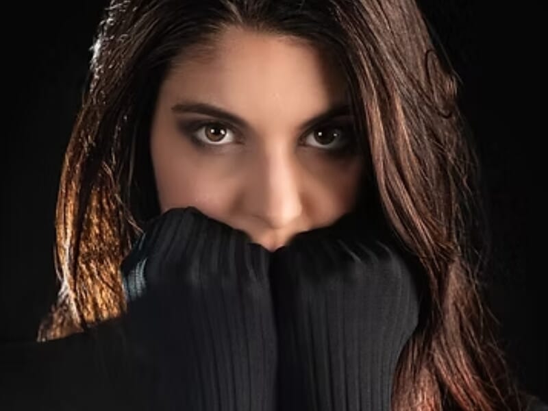 Femme aux longs cheveux bruns, pull noir couvrant la bouche, regard intense lors d'un shooting photo portrait près de Nancy. 