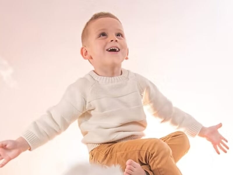 Garçon souriant en pull blanc et pantalon beige, assis avec les bras ouverts lors d'un shooting photo portrait près de Nancy. 