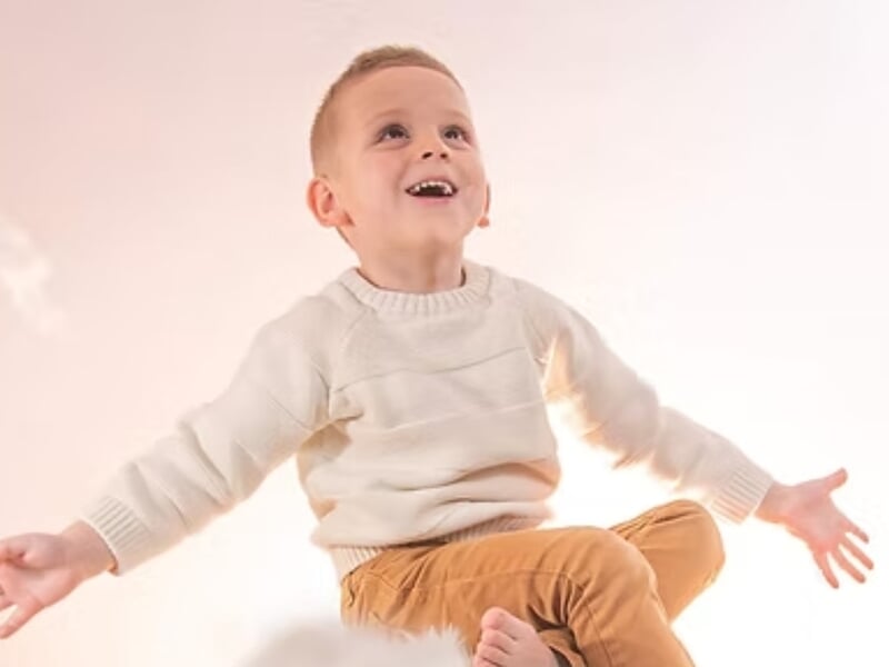 Garçon souriant en pull blanc et pantalon beige, assis avec les bras ouverts lors d'un shooting photo portrait près de Nancy.