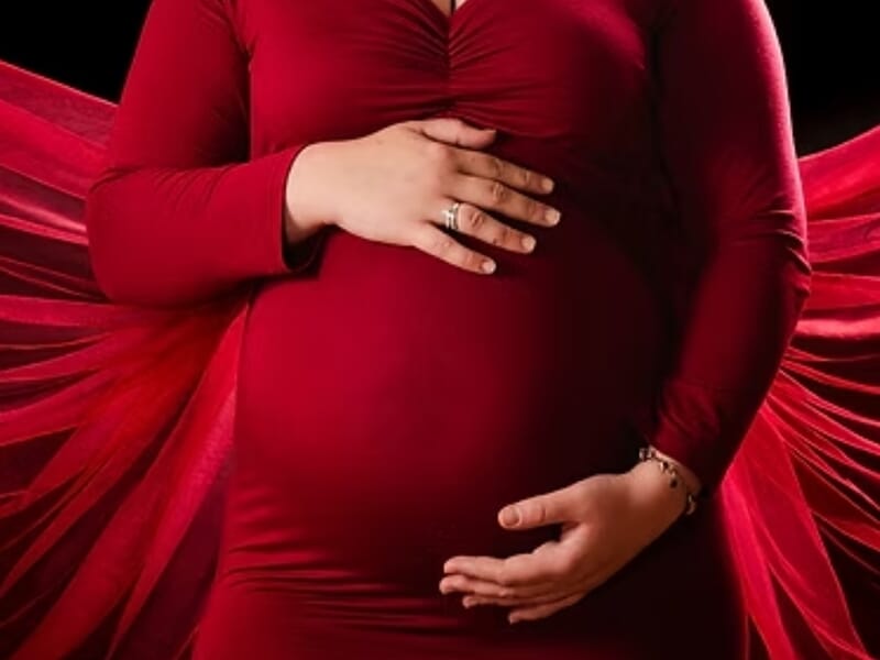 Femme enceinte en robe rouge tenant son ventre, avec un tissu rouge en arrière-plan lors d'un shooting photo grossesse près de Nancy.