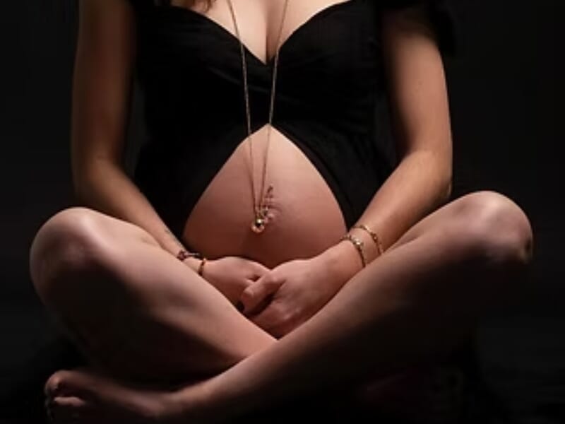 Femme enceinte assise en tailleur, vêtue de noir, mains posées sur son ventre lors d'un shooting photo grossesse près de Nancy. 