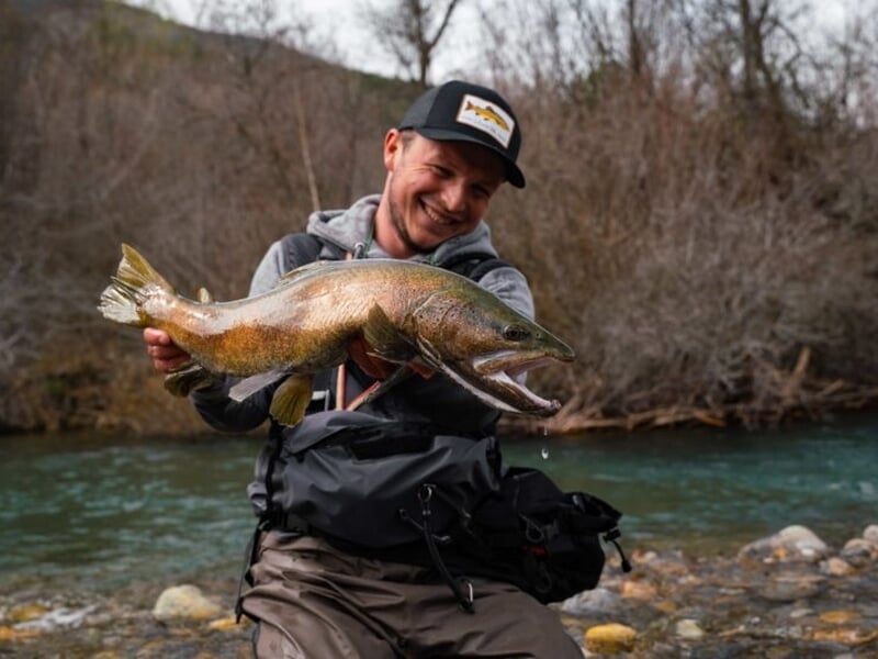 Homme souriant tenant un gros poisson pêché au bord d'une rivière en pleine nature, lors d'un stage de pêche au Lac de Caillauas.