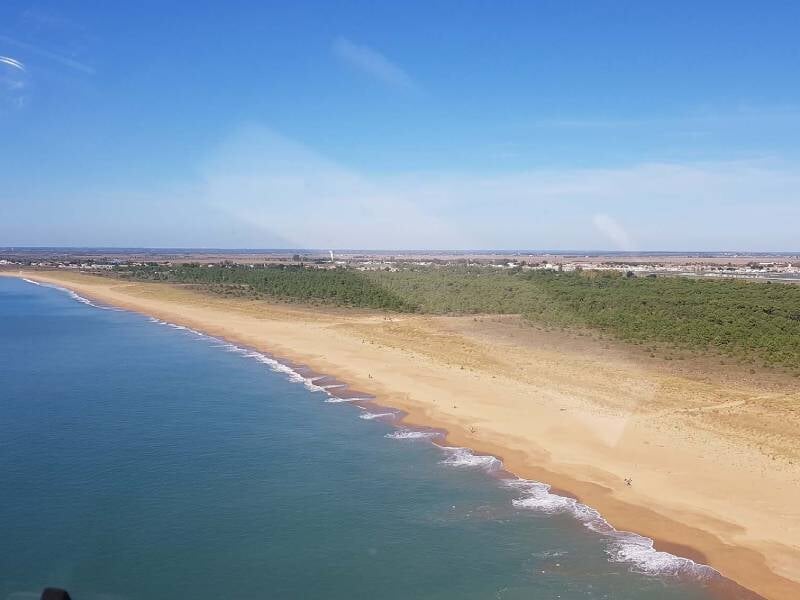 Vue aérienne d'une longue plage de sable fin bordée par une mer calme sous un ciel bleu clair.