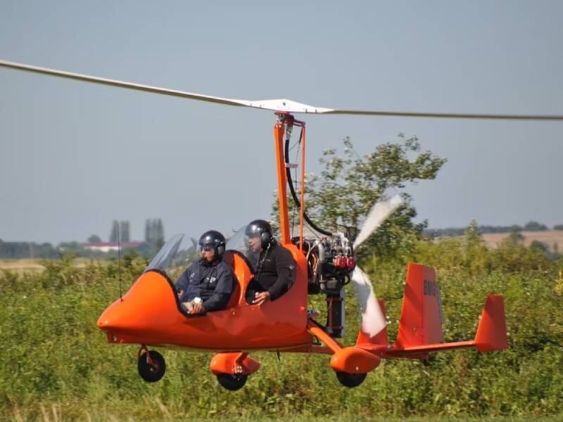 ULM autogire orange avec deux personnes à bord volant bas au-dessus d'un champ vert sous un ciel clair, lors d'un baptême de l'air dans le Marais poitevin.