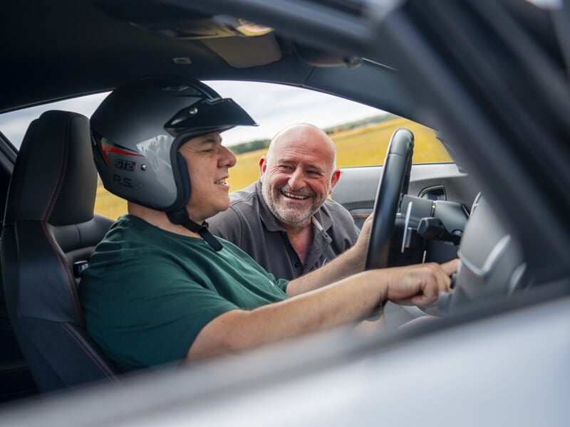 Homme avec casque souriant à un instructeur dans une voiture de sport lors d'un coaching de pilotage sur circuit.